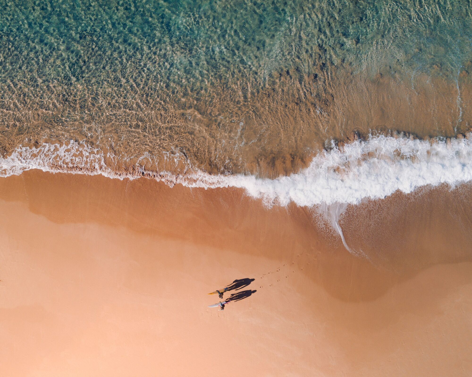 Aerial view of two surfers walking across golden sand beside turquoise surf